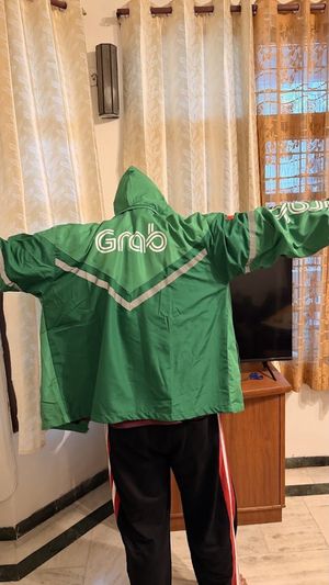 Kanishk seen from behind with his arms stretched out, wearing a green Grab delivery rain jacket several sizes too big, in a living room