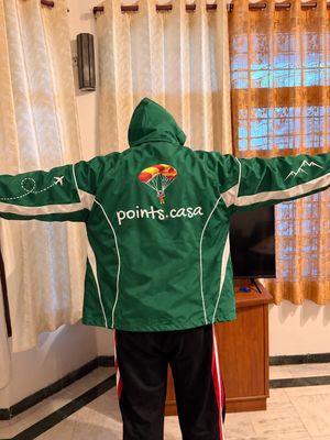 Kanishk seen from behind with his arms stretched out, wearing a green Grab delivery rain jacket several sizes too big, in a living room